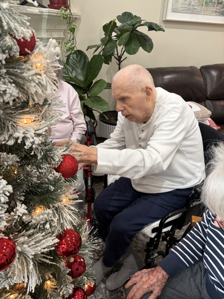 Older man seated beside a Christmas tree as he hangs a red ornament.