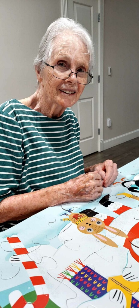 Older woman smiling while working on a colorful jigsaw puzzle at a table.
